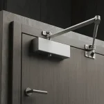 Un ferme-porte argenté et élégant à montage en surface avec un bras mécanique est installé en haut d'une porte moderne à texture bois, intégrée dans un mur en béton.