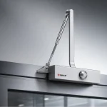 A imagem mostra um close-up de uma fechadura de porta hidráulica da marca OUBAO prateada montada na moldura superior de uma porta de vidro metálica moderna.
