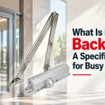 Silver door closer with folding arm mounted beside a glass entrance door, with text reading What Is Door Closer Backcheck? A Specification Guide for Busy Entrances.